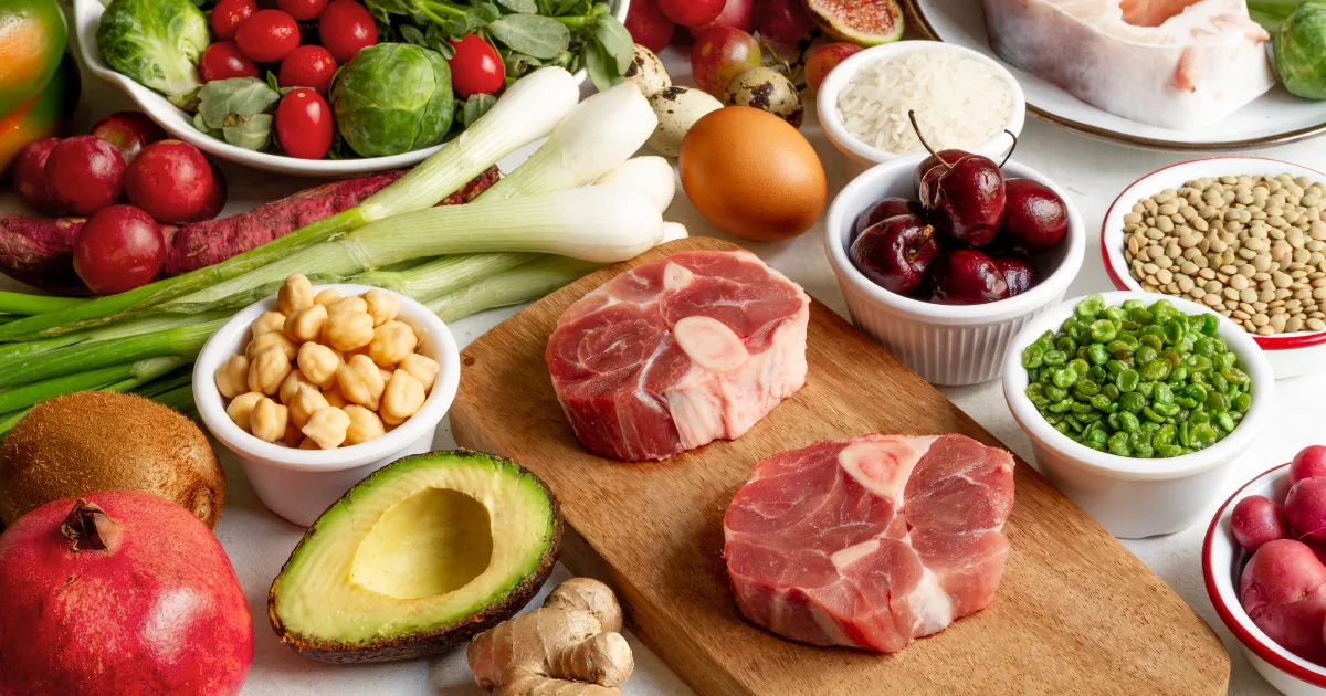 Fresh whole foods including raw beef, avocado, vegetables, legumes, nuts, and greens arranged on a counter top.