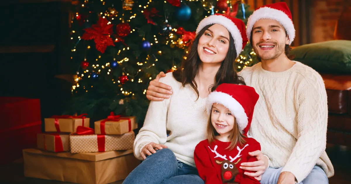 Young family of three posing over Christmas tree background, daughter holding an xmas gift