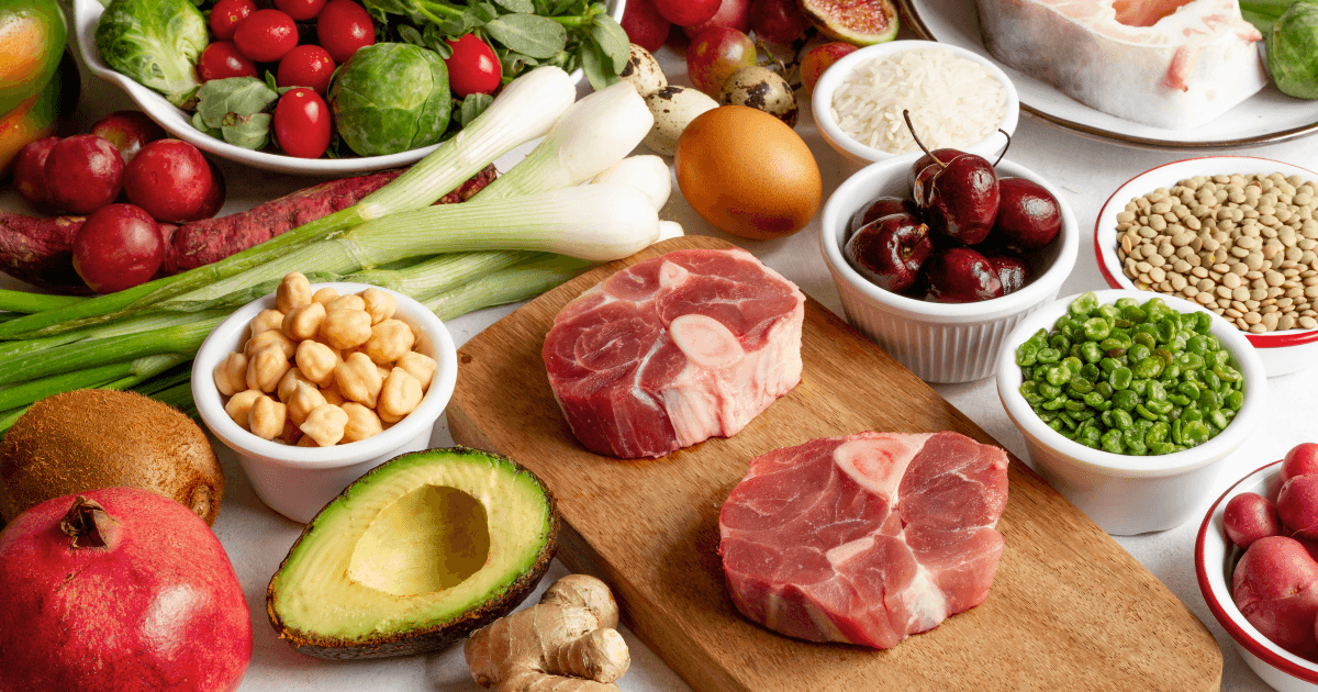 Fresh whole foods including raw beef, avocado, vegetables, legumes, nuts, and greens arranged on a counter top.
