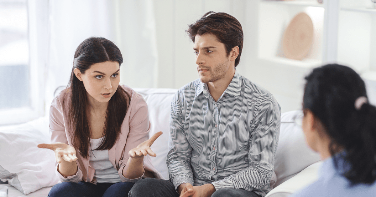 Woman in pink cardigan gesturing while speaking to a man in striped shirt during what appears to be a counselling session.