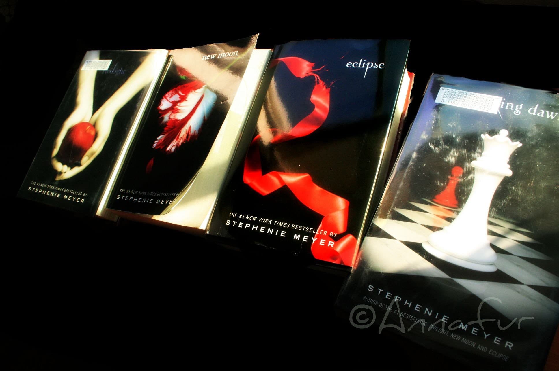 The four Twilight saga books by Stephenie Meyer arranged on a dark surface, showing their distinctive dark covers with red and white imagery.