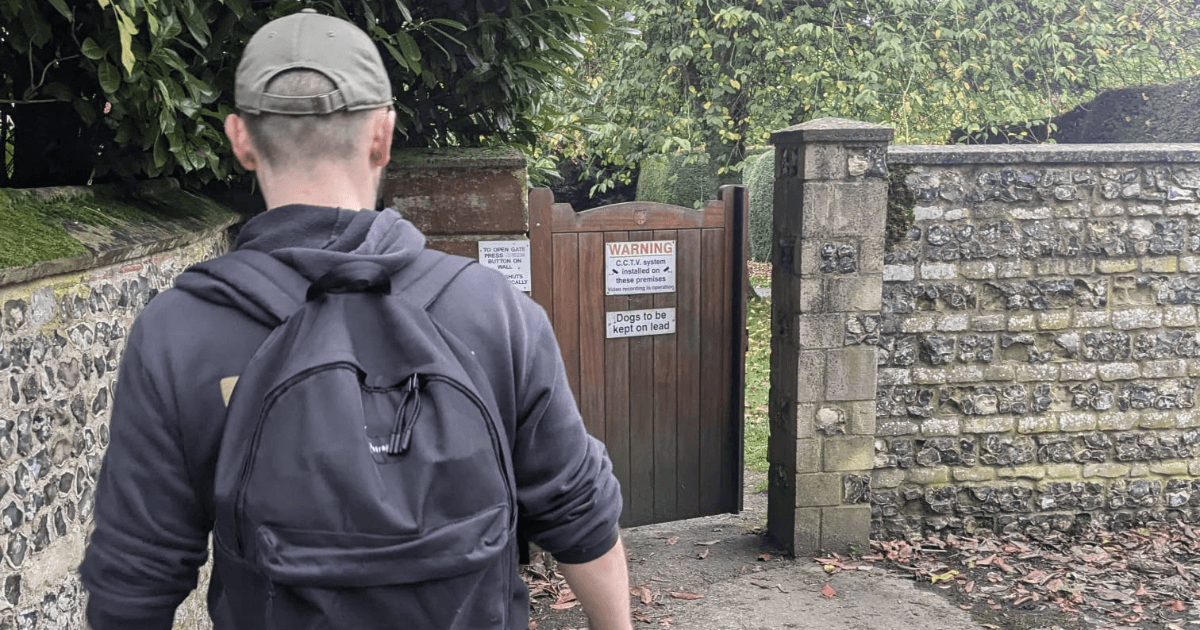 Person wearing a black hoodie and backpack walking towards a wooden gate with warning signs, viewed from the back.